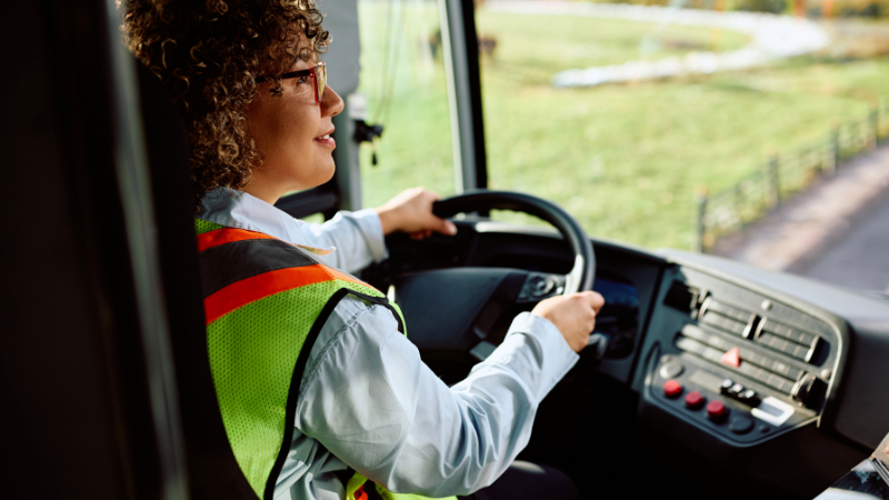A person wearing a safety vest and glasses is driving a bus, looking forward with hands on the steering wheel. The view outside the window shows greenery and a winding path.