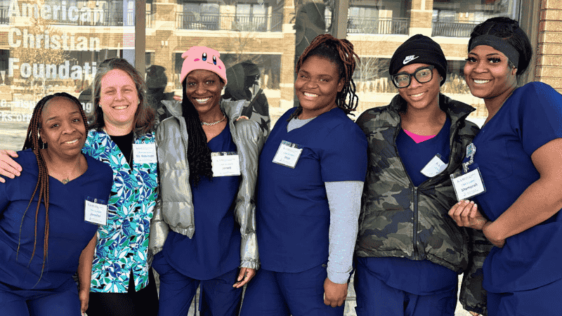 Six women stand together smiling outdoors, wearing blue medical scrubs and name tags. One woman wears a black headscarf, another a pink hat, and one wears glasses with a black beanie. A sign reads American Christian Founda….
