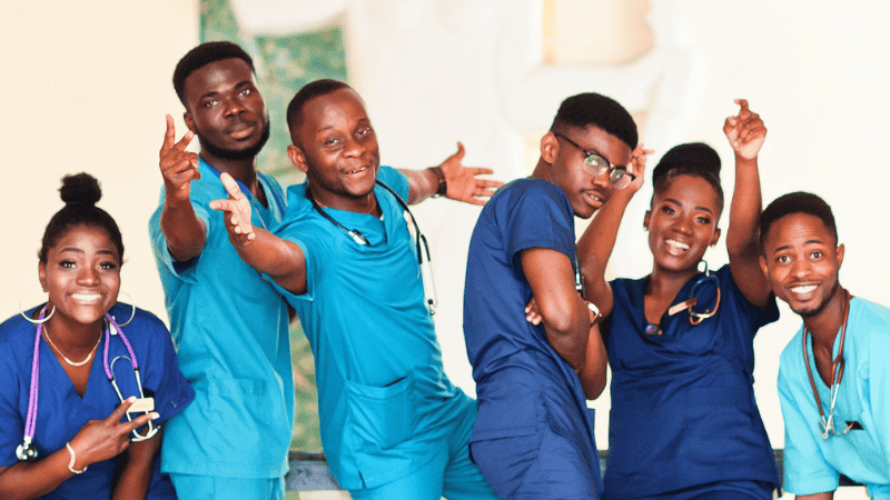 Six smiling healthcare professionals in scrubs pose playfully together, some with stethoscopes around their necks, against a light background.