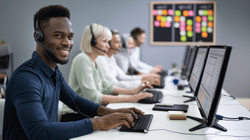 A group of customer service representatives wearing headsets work at computers in an office. The person closest to the camera is smiling and looking at the camera, while others focus on their screens.
