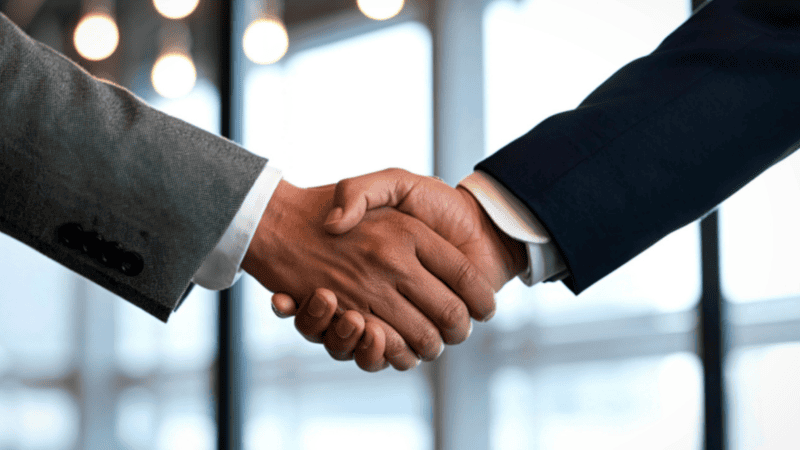 Two people in business attire shaking hands, symbolizing agreement or partnership, with blurred lights and windows in the background.