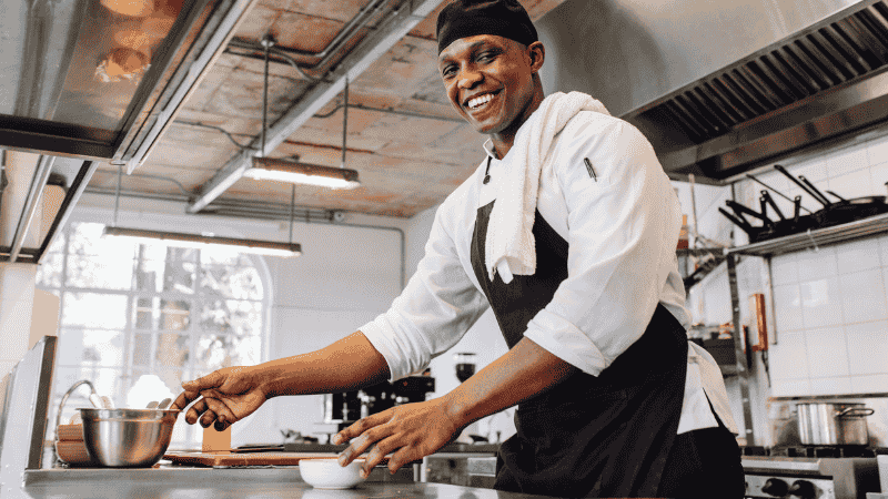 A smiling chef in a white uniform and black apron stands in a commercial kitchen, holding a spoon over a bowl, with a towel draped over his shoulder and cookware visible in the background.
