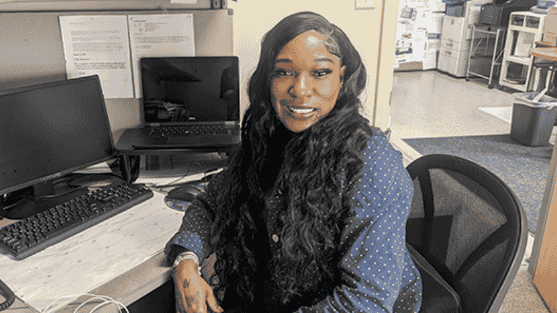 A woman with long, wavy hair sits at a desk in an office, smiling at the camera. She is wearing a blue polka-dotted shirt and is surrounded by computer monitors and office equipment.