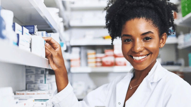 A pharmacist wearing a white coat smiles while reaching for a box on a shelf filled with medication in a pharmacy.