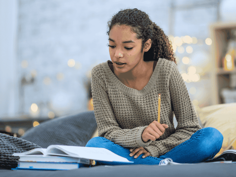 A young woman with curly hair sits cross-legged on a bed, wearing a sweater and jeans, studying an open notebook with a pencil in her hand. She appears focused, with books and papers around her.