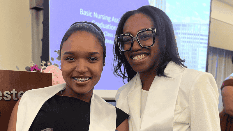 Two women wearing white sashes smile at the camera. One has braces and a headband; the other wears large glasses. A projector screen behind them displays text about a Basic Nursing Assistant graduation.