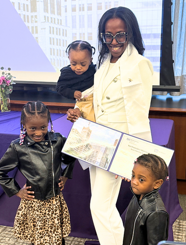 A woman with long, wavy hair sits at a desk in an office, smiling at the camera. She is wearing a blue polka-dotted shirt and is surrounded by computer monitors and office equipment.