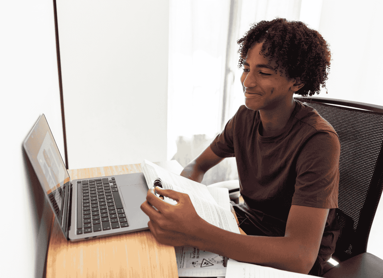 Teenager sitting at a desk, smiling while looking at a laptop screen, holding an open book and pen, appearing to study or participate in an online class.