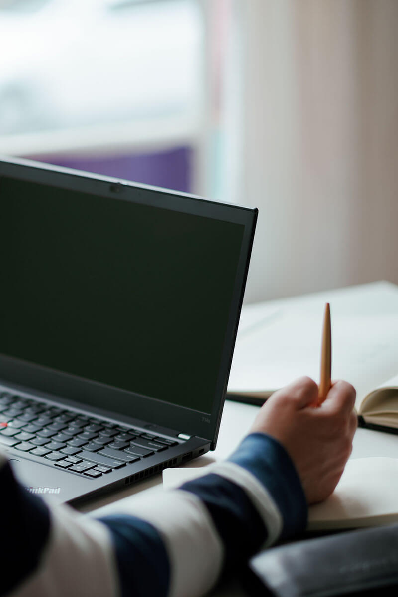 A person holding a pen sits at a desk with an open laptop and a notebook, appearing to be working or studying. The scene is indoors with soft, natural light.