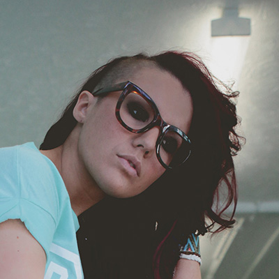 A person with long, dark, partially shaved hair and large, round glasses looks at the camera. They are wearing a light blue shirt and bracelets, with a light source visible in the background.